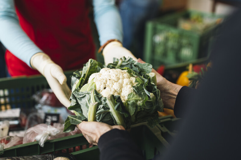 Zwei Personen tauschen Blumenkohl an einem Verkaufsstand aus. Eine Person trägt Handschuhe. Im Hintergrund sind grüne Kisten mit weiteren Lebensmitteln zu sehen.