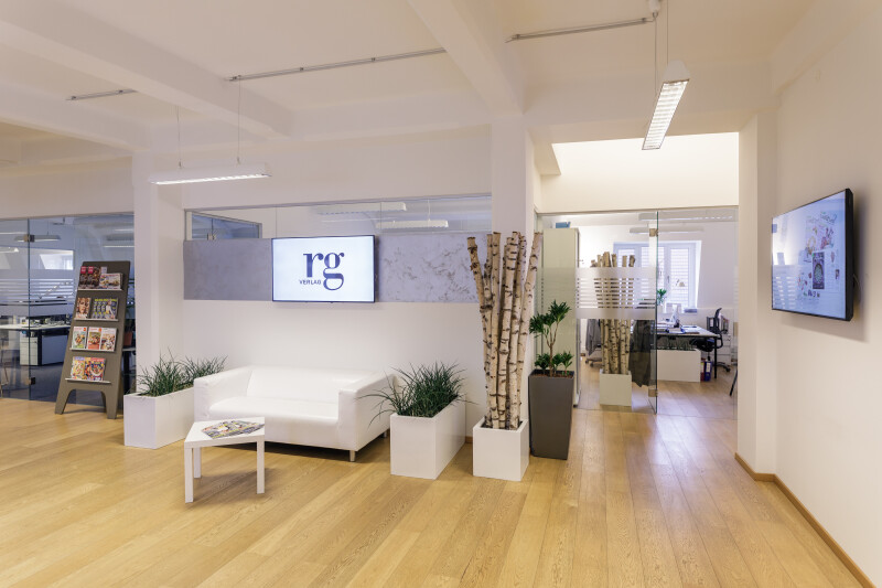 Modernes Büro mit Holzboden und weißen Wänden. Ein weißes Sofa steht neben einem kleinen Tisch mit Pflanzen daneben. Im Hintergrund sind Glaswände und ein Arbeitsbereich sichtbar. Zwei Bildschirme an den Wänden zeigen Inhalte. Helle Beleuchtung sorgt für eine freundliche Atmosphäre.