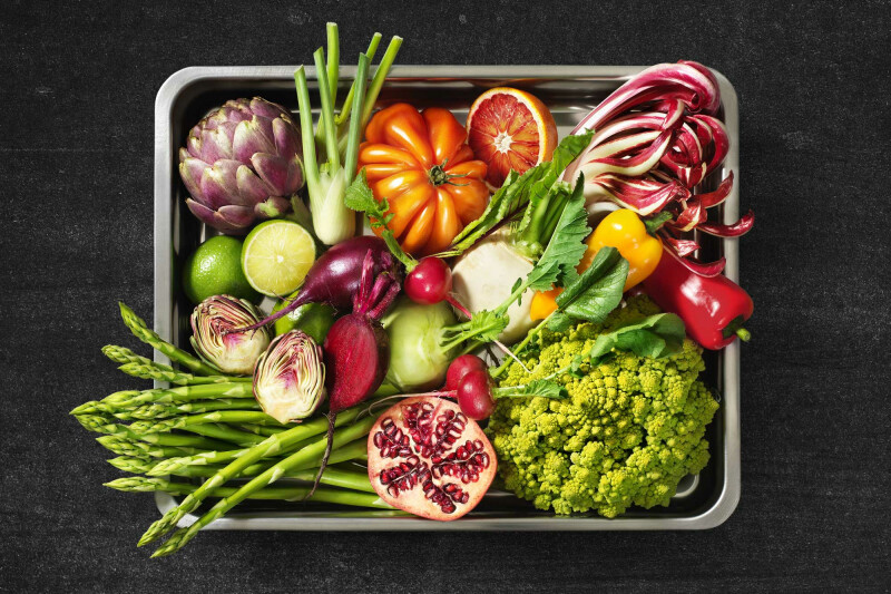 Ein Tablett mit einer Vielzahl von frischem Gemüse und Obst auf schwarzem Hintergrund. Darunter befinden sich Artischocken, Spargel, bunte Paprika, Romesco, Radieschen, halbierte Limetten, eine halbierte Blutorange und eine Granatapfelscheibe.