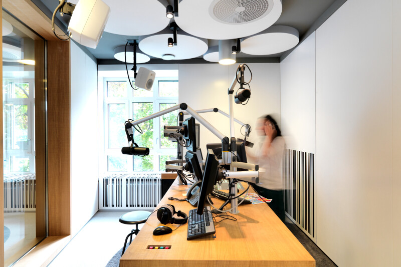 Moderner Radiostudio-Raum mit Schreibtischen, Mikrofonen und Computern. Eine Person steht unscharf neben den Arbeitsplätzen. Große Fenster lassen Tageslicht herein. Decke mit runden Akustikelementen.