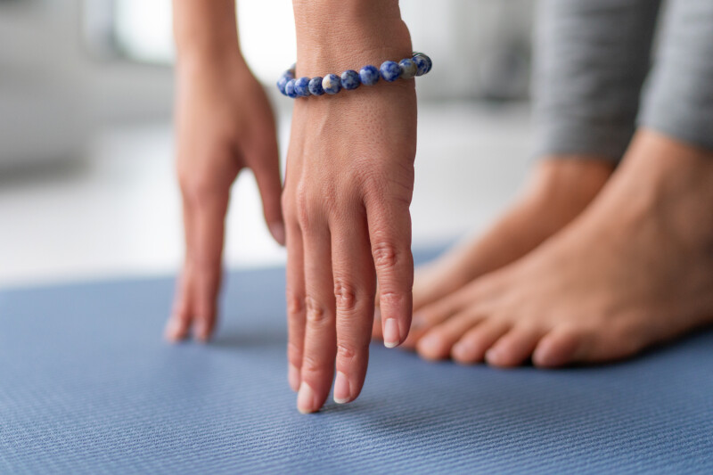 Hands reaching towards feet in a forward bend position on a blue yoga mat. A person is wearing a beaded bracelet.