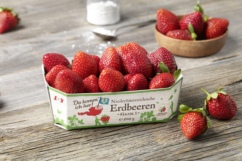A cardboard container filled with fresh strawberries placed on a rustic wooden surface. Additional strawberries are scattered around and a small bowl of strawberries is in the background. The container label reads in German, indicating they are Austrian strawberries.