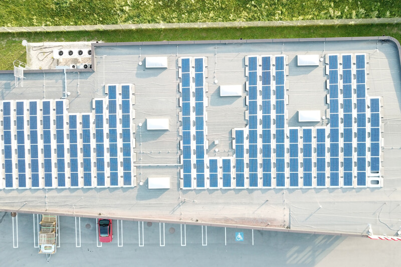 Aerial view of a flat rooftop covered with evenly spaced solar panels, with a grassy area and parked cars adjacent to the building.