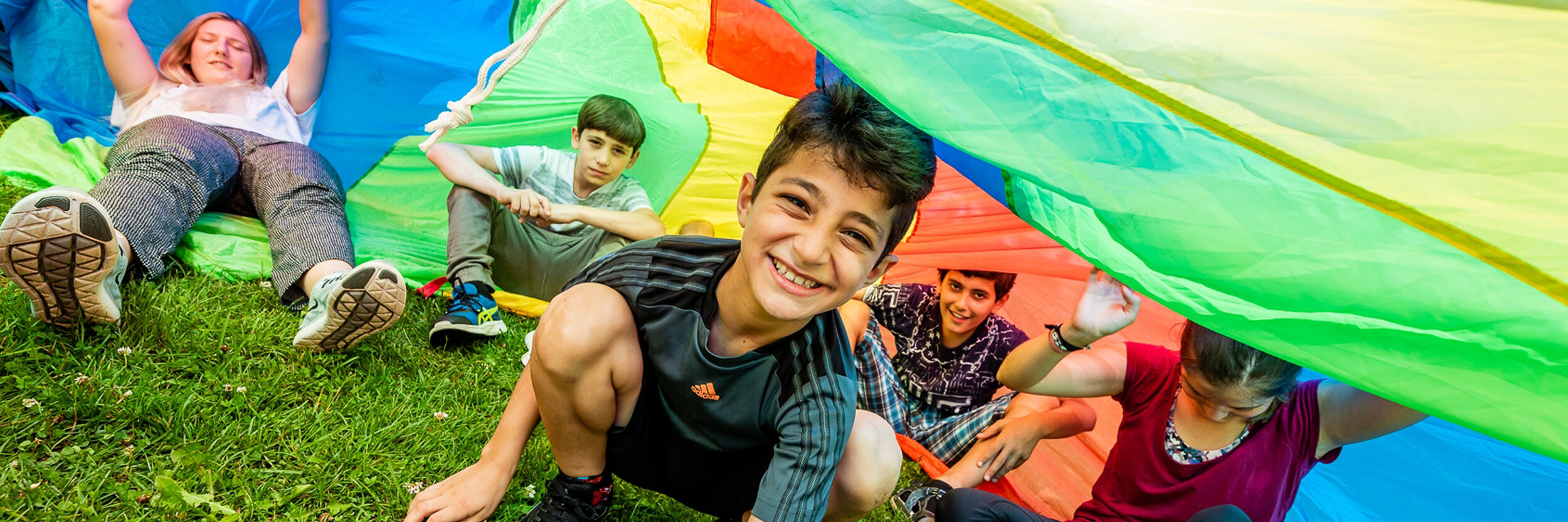 Children smiling and playing under a colorful parachute on a grassy area.