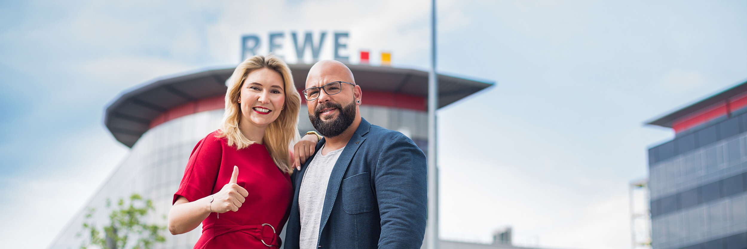 A smiling woman in a red dress giving a thumbs-up stands next to a bald man with glasses, wearing a dark jacket. They are outdoors, with a large building displaying the word "REWE" in the background.