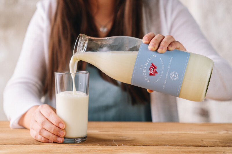 A person pouring milk from a glass bottle with a blue label into a clear drinking glass on a wooden table.