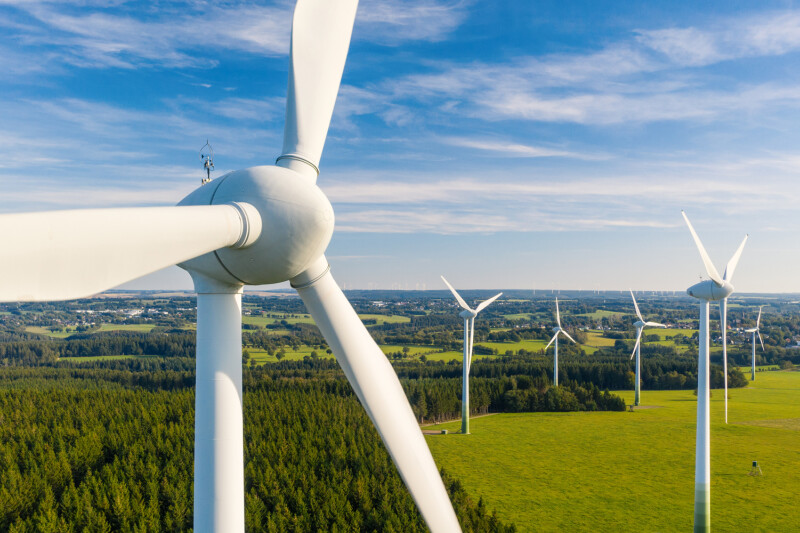 Aerial view of several wind turbines in a green field with a forest in the background, under a blue sky with scattered clouds.