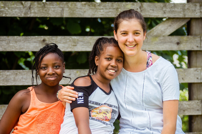 Three people are smiling and sitting together in front of a wooden fence. Two young girls are on the left, and an adult woman is on the right with her arm around one of the girls. They appear relaxed and happy.