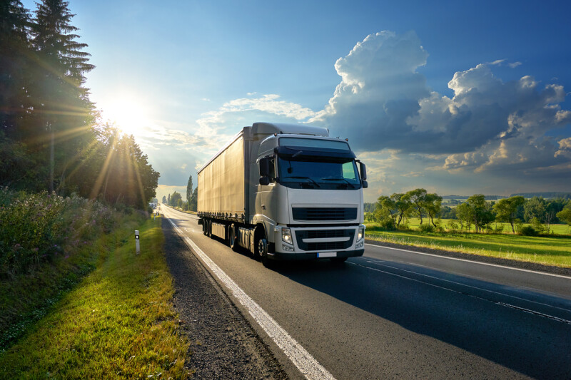 A white semi-truck driving on a rural road with lush greenery, trees, and bright sunlight filtering through clouds in the sky.