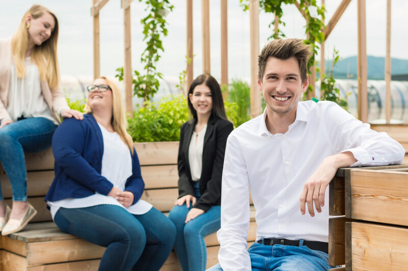 Four people smiling and sitting on wooden planters in a rooftop garden. One person is in the foreground wearing a white shirt, while three others are seated together in the background surrounded by greenery.