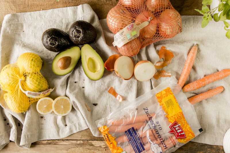 Various groceries are spread out on a wooden table and linen cloth, including whole and halved avocados, netted lemons, sliced lemons, a bag of sweet onions with loose onions and peel, several carrots, and a bag of sugar. Fresh herbs are visible in the corner.