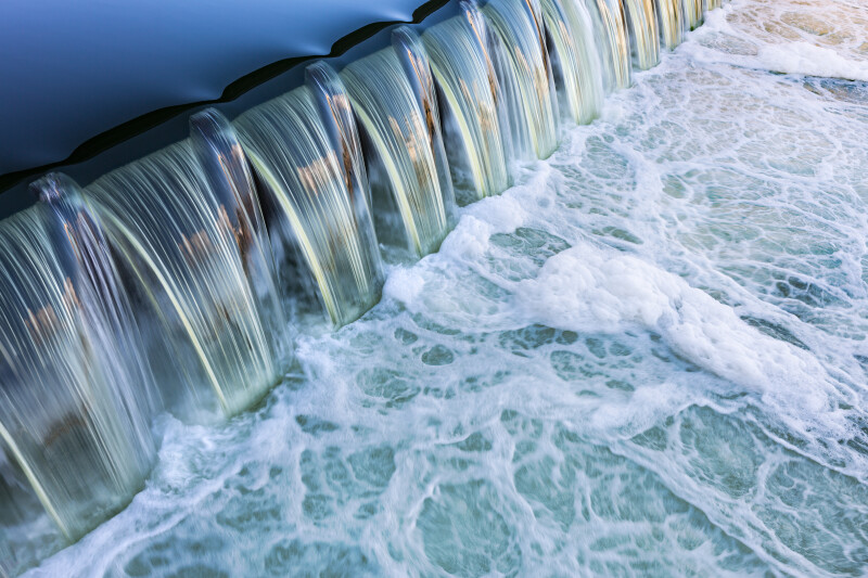 Water cascading over a small dam, creating a fluid, wavy pattern with frothy white water below.
