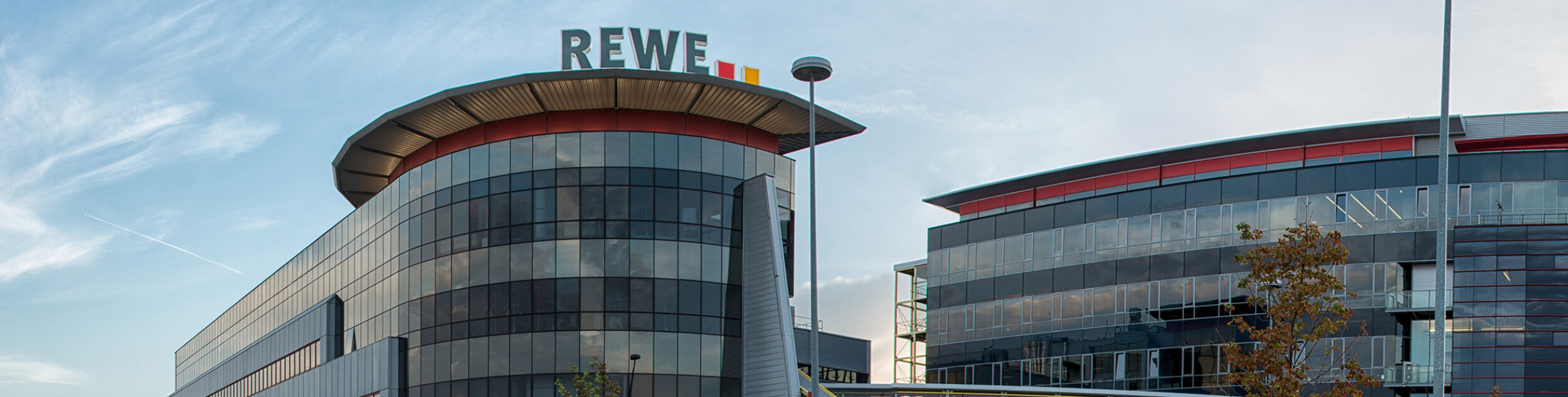Modern office buildings with glass facades, featuring the REWE logo on top of the left building. A clear sky is visible in the background.