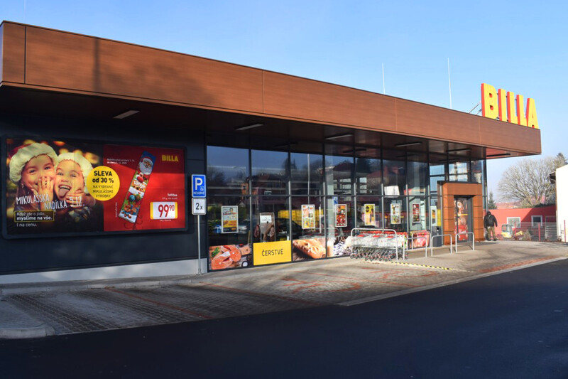 Exterior of a Billa supermarket with a modern glass facade reflecting sunlight. A large promotional sign displays images of food and discount information in Czech. Shopping carts are lined up near the entrance. One person is walking towards the entrance.