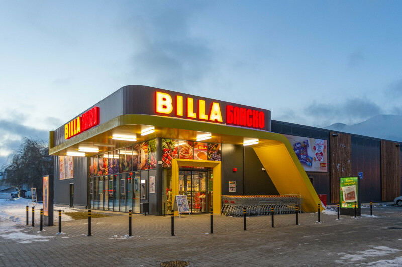 A modern supermarket with bright red "Billa Bansko" signage on the exterior. The building features large windows displaying various product images. The entrance is illuminated, and a snowy landscape surrounds the store.