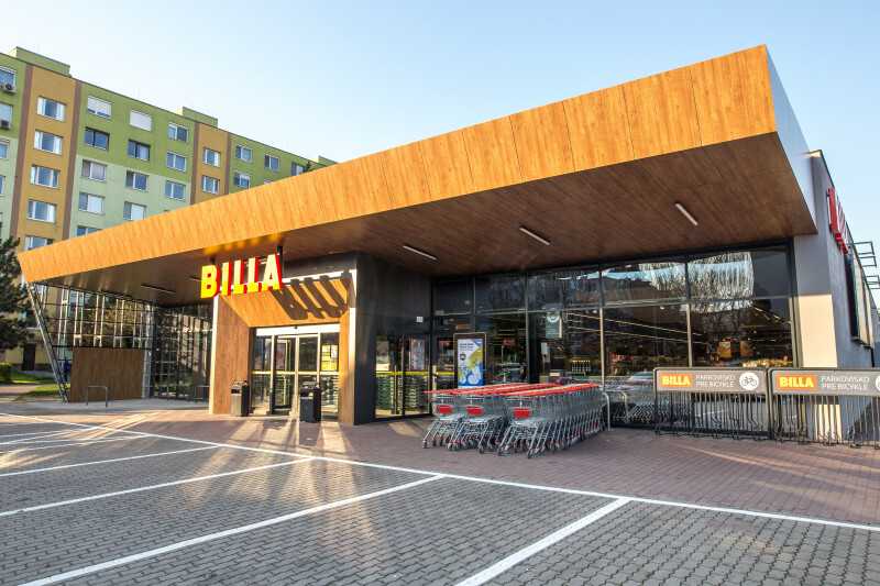 The exterior of a Billa supermarket with a modern wooden facade. A row of shopping carts is lined up near the entrance, and an empty parking lot is in the foreground. A residential building is visible in the background under a clear sky.