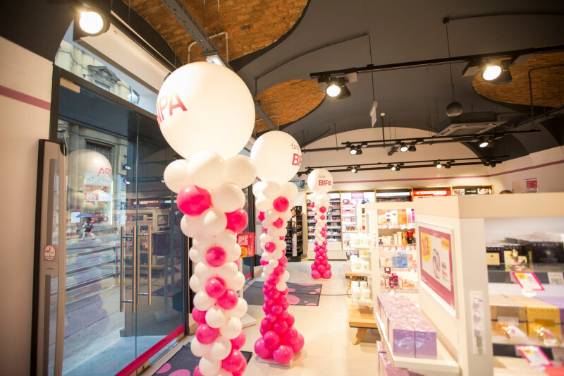 A brightly lit cosmetic store interior featuring columns of pink and white balloons with "BIPA" printed on them. The store has high ceilings with exposed brick arches and shelved displays of beauty products. Large storefront windows reveal a street outside.