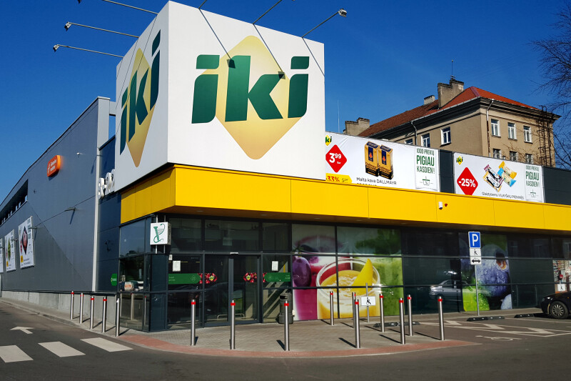 A modern supermarket exterior with the "iki" logo prominently displayed on a large sign. The building features colorful promotional banners and a parking area with visible bollards. Nearby, a few residential buildings are partially visible under a clear blue sky.