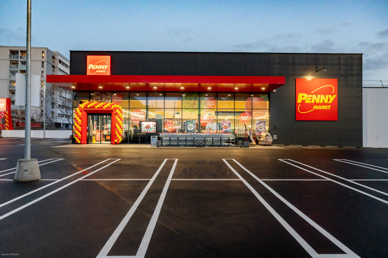 A Penny Markt store with a bright red and yellow entrance, located in a parking lot with clearly marked parking spaces. The store has large windows displaying promotional posters.