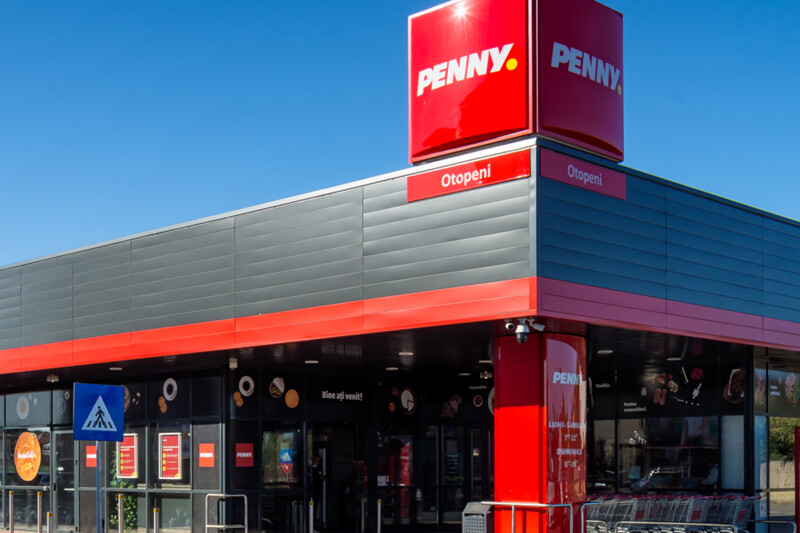 A modern Penny supermarket with red and grey exterior features, located in Otopeni. Bright signage and a clear blue sky in the background.