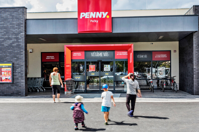 People walking towards the entrance of a Penny supermarket. The entrance is a bright red color with glass doors. Shopping carts are visible on the left, and bicycles are parked on the right. A sign above the door reads "Vítáme vás."