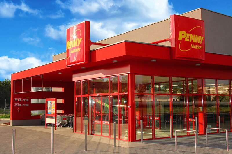 A bright red Penny Market building with large glass windows and doors. Sunny day with a partly cloudy sky. Shopping carts are visible near the entrance.