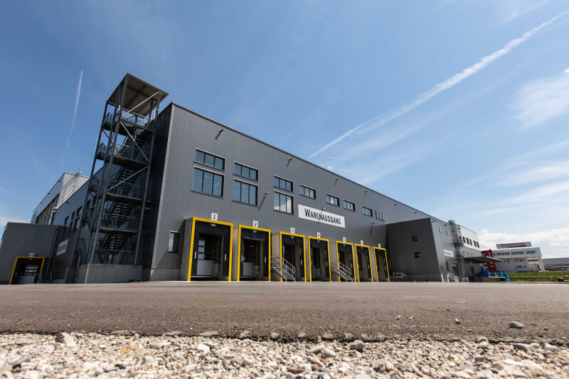 Large industrial warehouse with multiple loading docks, yellow accents, and a clear blue sky overhead. The foreground features a paved area and gravel along the edge.