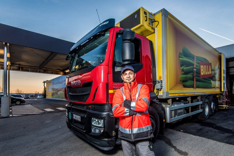A man in an orange safety jacket and cap stands confidently with arms crossed in front of a large red and yellow box truck parked under a shelter. The truck has images of vegetables on its side and a brand logo. The sky is clear, indicating a late afternoon or early evening.